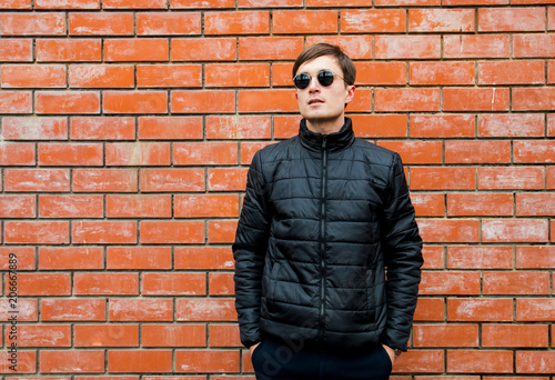 a young man stands leaning against a brick wall in sunglasses cross his arms