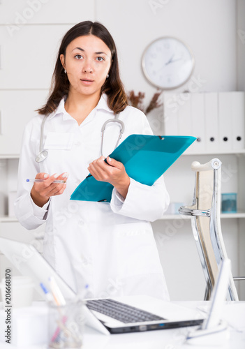 Girl doctor in white medical coat holding clipboard in office