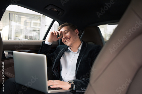 Be always cheerful and positive! Cheerful acknowledged young professional businessman sitting in the car and smiling, while working on the laptop. © stacestock