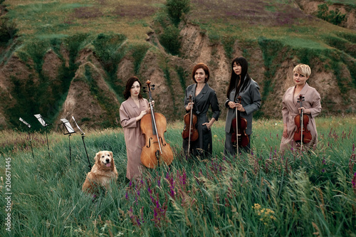 Tablou pe pânză Female musical quartet with three violins and one cello prepares to play at flowering meadow against backdrop of picturesque landscape next to dog