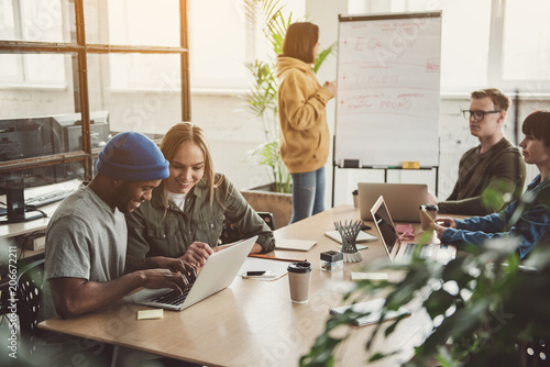 Beaming girl and cheerful male looking at laptop while having job with partners in office. Female describing information while locating near board. Start up team concept
