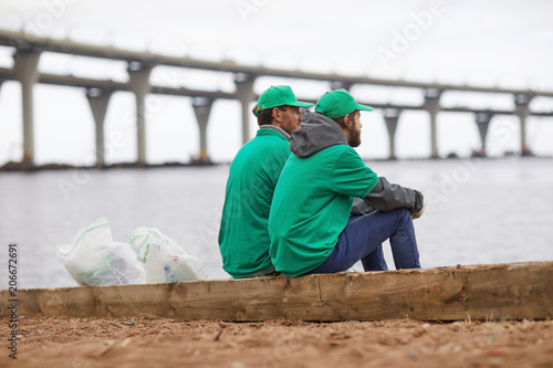 Two guys in greenpeace uniform sitting on river bank by water with sacks full of litter near by