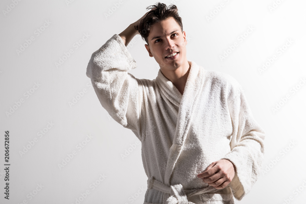 Ready to new day. Portrait of cheerful cute young man in white terrycloth bathrobe is looking forward with smile. He is standing against light wall with raised hand. Male beauty concept. Copy space