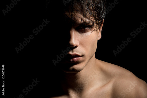 Close up portrait of young mysterious guy with half face in shadow. He is standing on dark background and expressing thoughtfulness. Masculine beauty concept