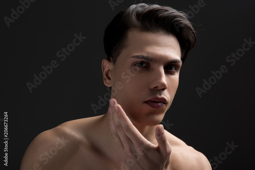 Portrait of gorgeous young shirtless man with perfect skin is applying moisturizing cream on his face. He is looking at camera confidently while standing against dark background. Male skincare concept