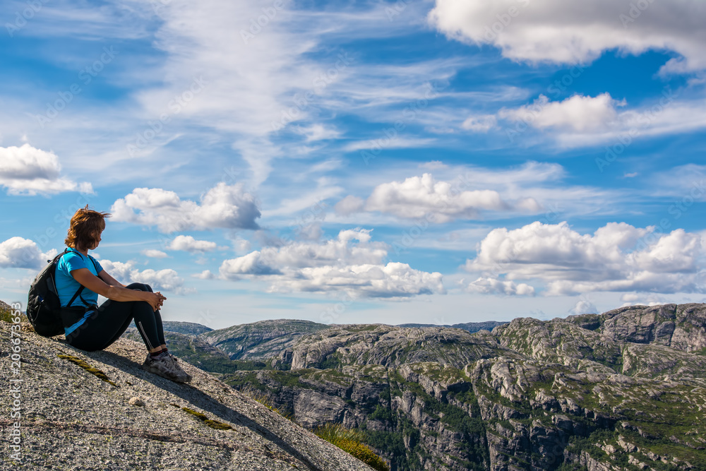 A woman is sitting on the edge of cliff on the way to boulder (Kjeragbolten) stuck in between the mountain crevices of Kjerag above Lysefjord, near Lysebotn, Norway. The feeling of complete freedom