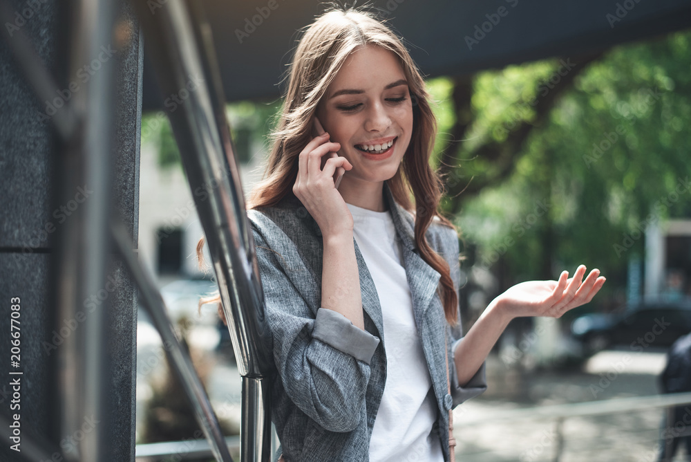 Waist up portrait of smiling young woman communicating on the smartphone. She is standing outside and leaning against railing