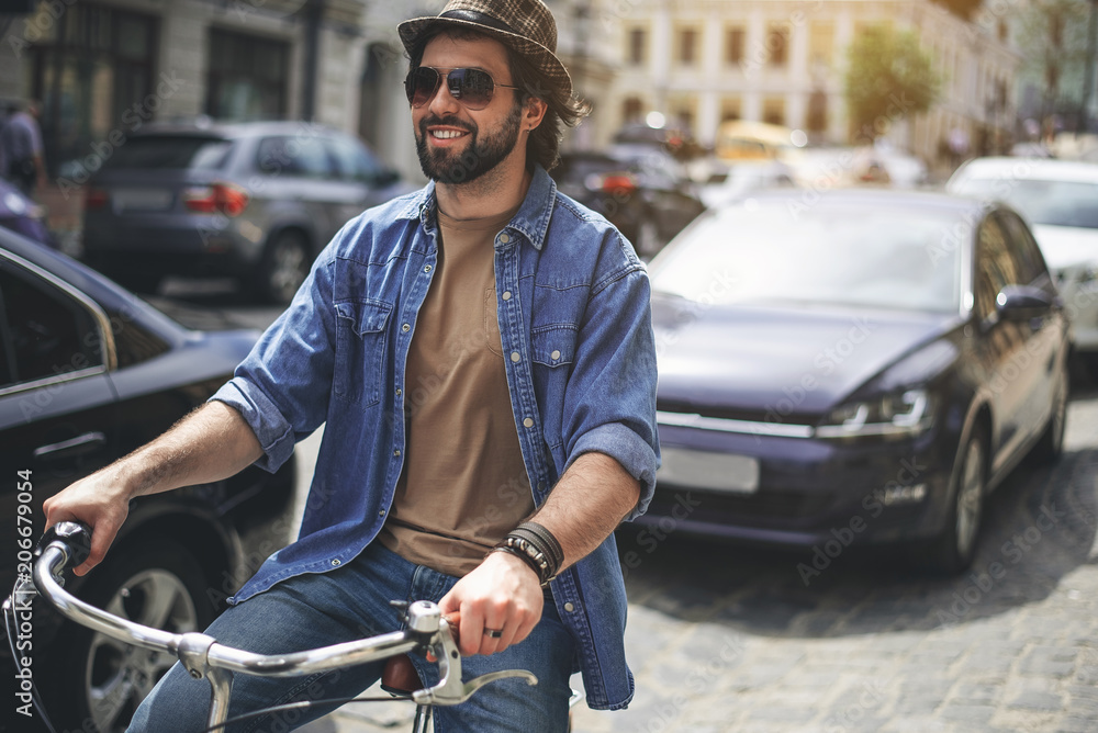 Life in movement. Portrait of cheerful man in headwear and sunglasses riding bike along the road. Cars on background. Copy space in right side Life in movement. Portrait of cheerful man in headwear and sunglasses riding bike along the road. Cars on background. Copy space in right side