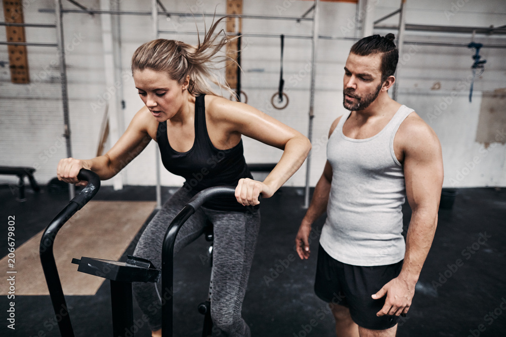 Young man watching is friend ride a gym stationary bike Young man watching is friend ride a gym stationary bike