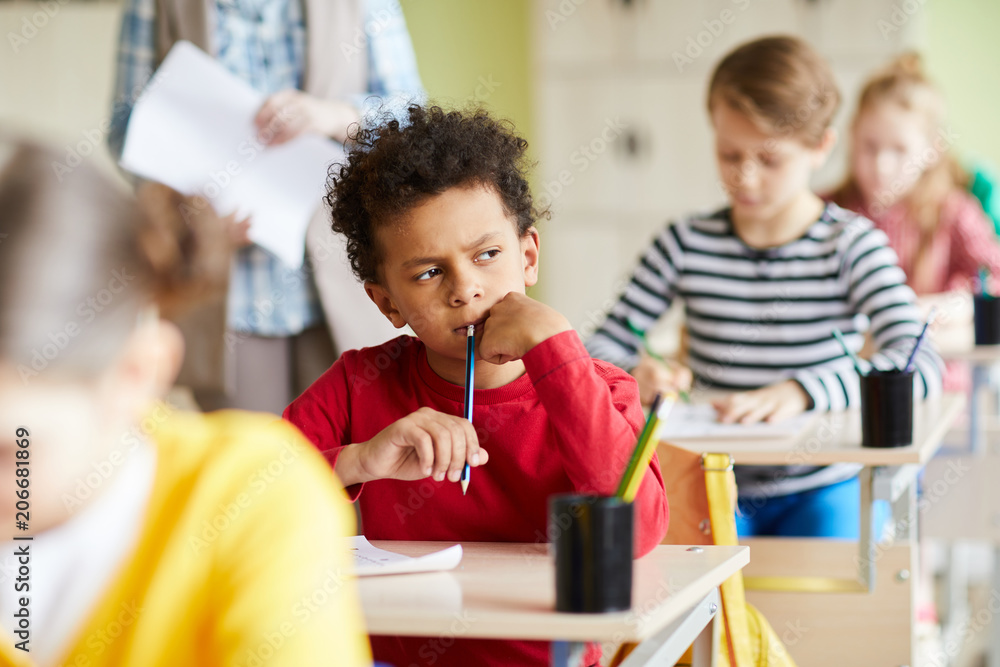 Obraz premium Pensive schoolboy sitting by individual desk and thinking of right answer while writing exam test
