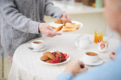 Mature female putting plate with bread slices on served table with desserts and fresh tea