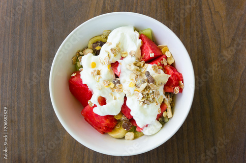 Delicious, healthy fruit salad bowl with muesli and yogurt for breakfast. Top view of dessert on wood background