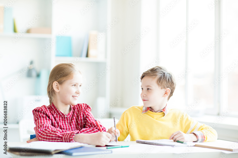 Two friendly little schoolchildren sitting by desk in classroom, thinking of ideas and discussing them Two friendly little schoolchildren sitting by desk in classroom, thinking of ideas and discussing them