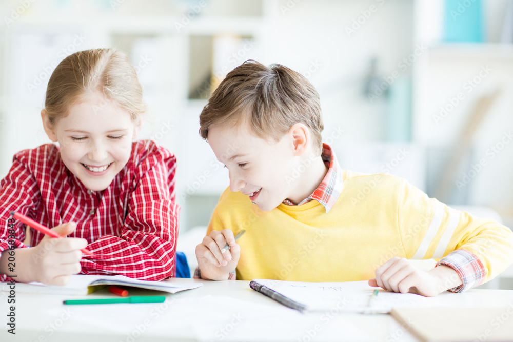 Two happy kids looking at picture in copybook of girl while sitting by desk at lessonn of drawing Two happy kids looking at picture in copybook of girl while sitting by desk at lessonn of drawing