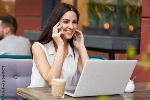 Portrait of pleasant looking female etrepreneur enjoys communication via cell phone, has pleasant smile, focused somewhere, uses modern laptop computer, spends free time at cozy coffee shop.