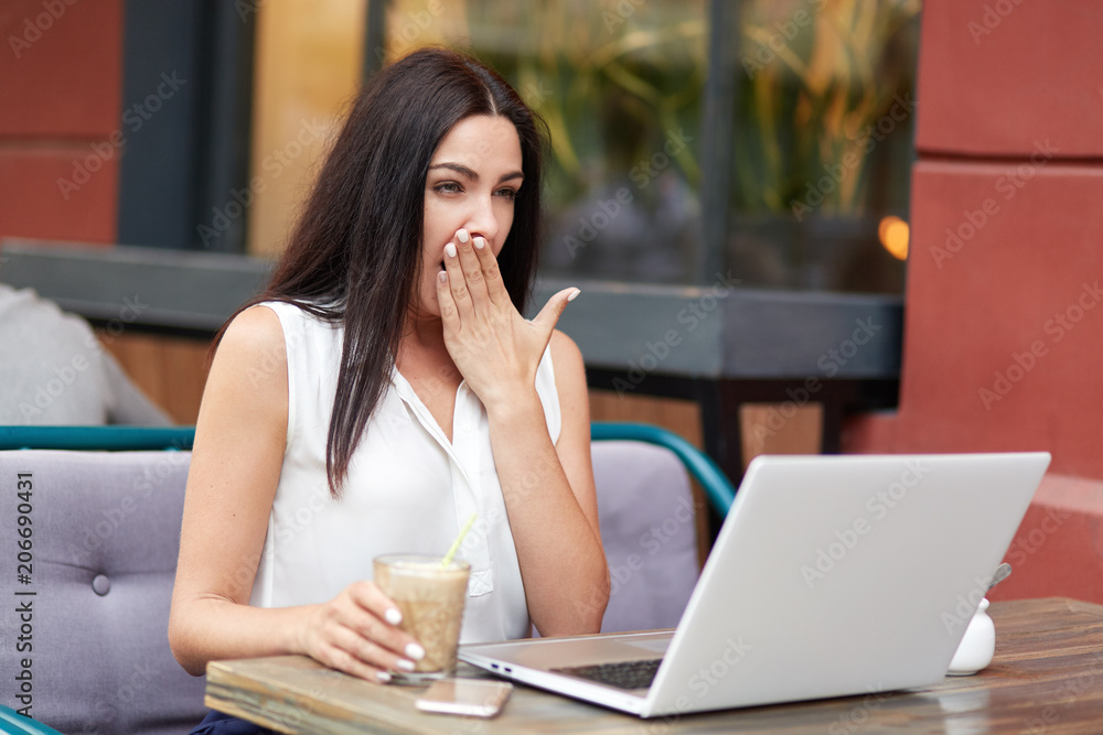 Tired overworked young female freelnacer yawns as want to sleep, wants to have rest, works long time at laptop computer, drinks cocktail, sits against cafe interior. People, tiredness concept Tired overworked young female freelnacer yawns as want to sleep, wants to have rest, works long time at laptop computer, drinks cocktail, sits against cafe interior. People, tiredness concept