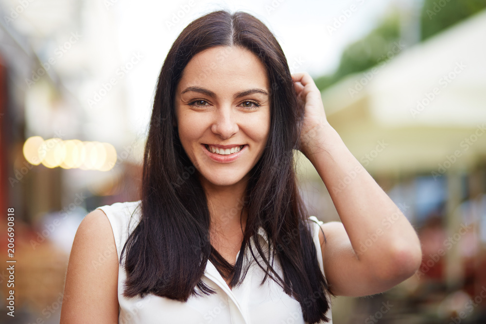 Close up portrait of young brunette female has pleasant smile, has healthy soft skin, stands against city blurred background. Cute woman being in high spirit. People, beauty, leisure concept