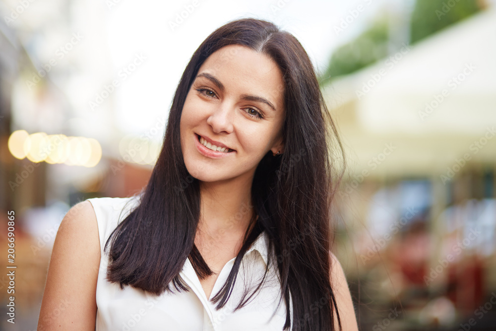Headshot of brunette female smiles joyfully, has healthy skin, poses against blurred background, enjoys stroll with her boyfriend. Portrait of good looking young woman shows her natural beauty