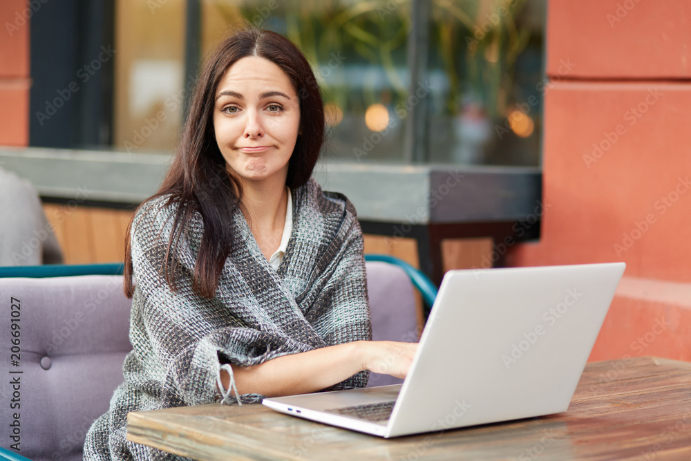 Puzzled tired female freelancer makes online research of information, wrapped in warm coverlet, poses against cafe interior. Attractive woman student studies online courses or going to watch webinar Puzzled tired female freelancer makes online research of information, wrapped in warm coverlet, poses against cafe interior. Attractive woman student studies online courses or going to watch webinar