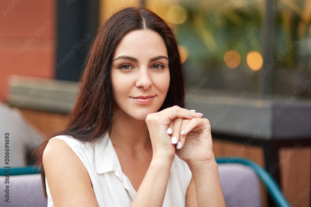 Close up shot of pretty brunette young female with dark hair, healthy skin, holds hands under chin, looks mysteriously at camera, has pleasant talk with friend, poses against blurred background Close up shot of pretty brunette young female with dark hair, healthy skin, holds hands under chin, looks mysteriously at camera, has pleasant talk with friend, poses against blurred background