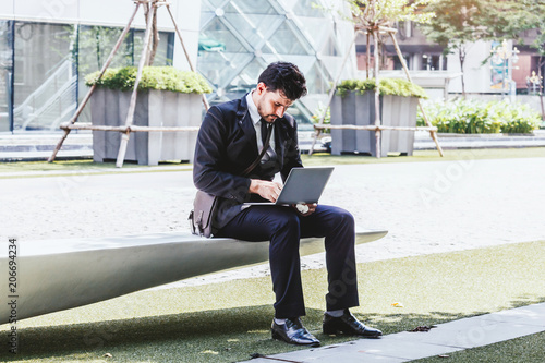 Handsome businessman in black elegant suit sitting and working with laptop computer on the city.Business and startup idea concept