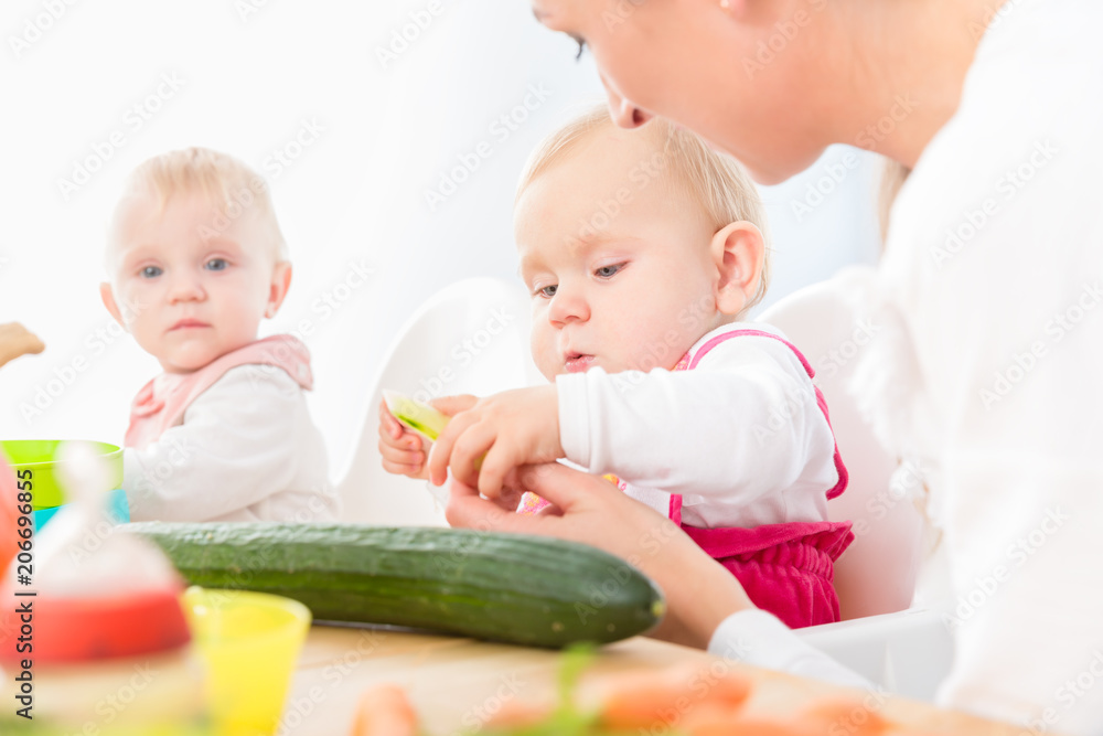 Portrait of a cute baby girl with blue eyes eating healthy solid food, while sitting in a high chair next to another baby in a modern daycare center for babies Portrait of a cute baby girl with blue eyes eating healthy solid food, while sitting in a high chair next to another baby in a modern daycare center for babies