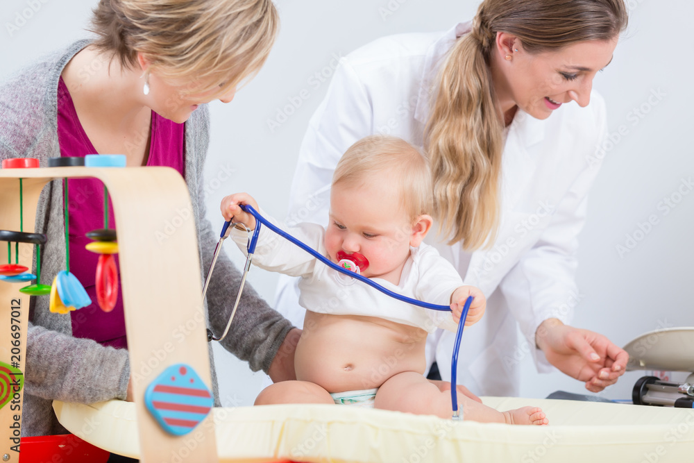 Cute and healthy baby girl playing with the stethoscope next to her careful mother, during routine medical check-up in a modern clinic with dedicated pediatricians Cute and healthy baby girl playing with the stethoscope next to her careful mother, during routine medical check-up in a modern clinic with dedicated pediatricians