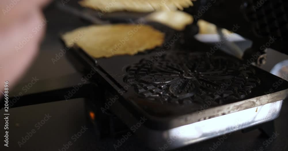A handheld shot of a woman opening a pizzelle maker to remove two freshly-cooked pizzelles and places new dough balls for the next batch. A handheld shot of a woman opening a pizzelle maker to remove two freshly-cooked pizzelles and places new dough balls for the next batch.
