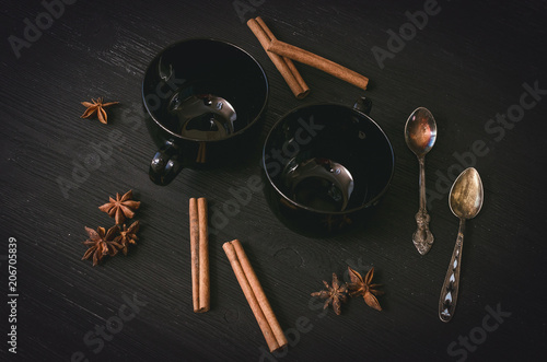 Two empty black tea cup and star anise with cinnamon and set of silver spoon around on black wooden table background.