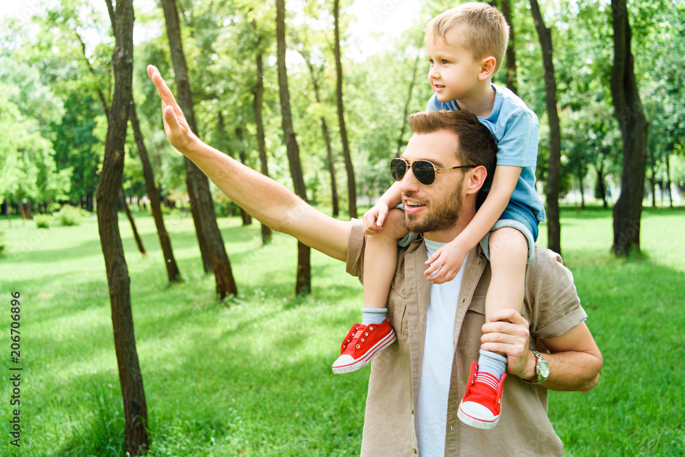 Obraz premium father waving hand to someone and holding son on shoulders at park