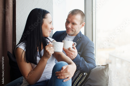 Young cute couple with a cup of hot coffee by the window smiling. Concept of love and relationship, family.