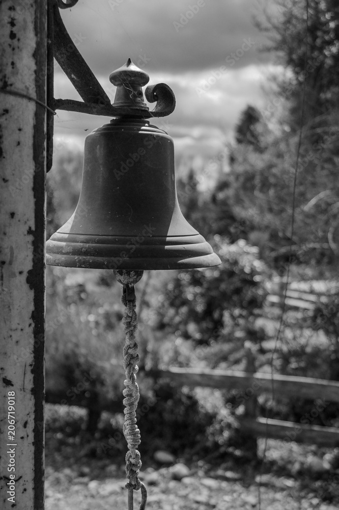 Obraz premium Bronze bell on a rusty post with a garden in the background in black and white.