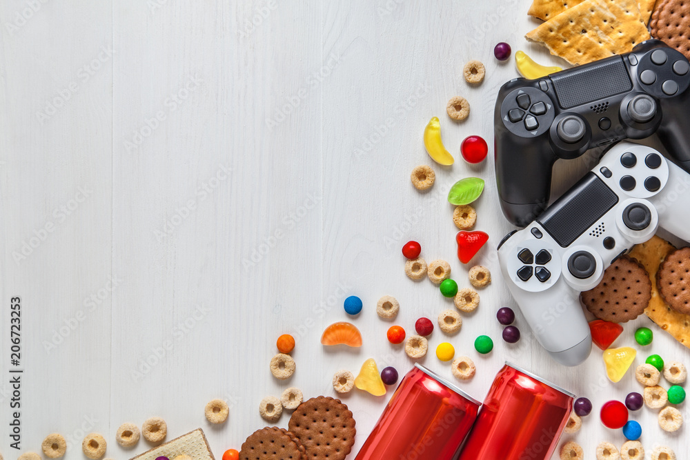 black and white modern gamepads on a white wooden background among sweets, biscuits and cans of soda. black and white modern gamepads on a white wooden background among sweets, biscuits and cans of soda.