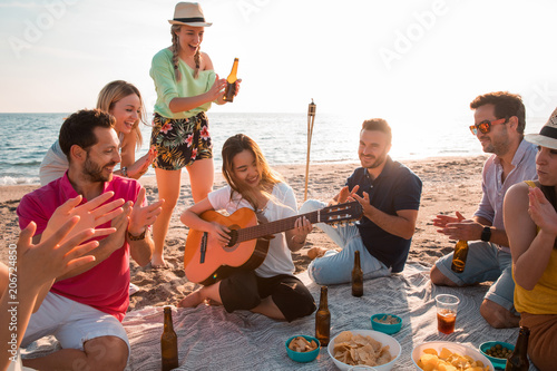 Happy multiracial young people playing guitar and making picnic at the beach. Friends are drinking beers and listening to music. Having fun at beach party in evening. photo