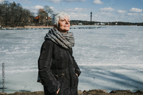 Cheerful middle-aged woman enjoying her holidays in Stockholm, Sweden. Walking around an icy lake on a sunny and cold spring day. Lifestyle photography.