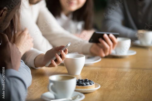 Diverse people using smartphones sitting at cafe table together, friends holding gadgets enjoying new apps or checking social networks at meeting connected online to public place wifi, close up view