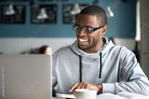 Smiling millennial african american casual man in glasses working with apps or communicating online on laptop sitting at cafe table, happy black businessman using public place wifi in coffee shop