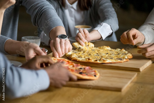 Hands of diverse people taking pizza slices from wooden board dining together, multiracial friends or colleagues sharing meal having lunch in cafe restaurant, italian pizzeria concept, close up view