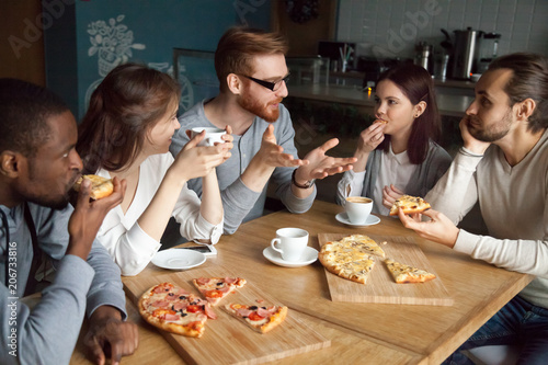 Redhead guy discussing interesting news with diverse friends hanging in cafe having lunch  millennial multiracial people students or colleagues eating pizza and talking together in pizzeria place