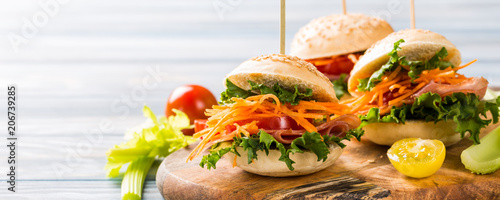 Homemade mini burgers with ham, tomato, carrot, fresh salad served on wooden board. Healthy junk food concept with copy space. Banner.