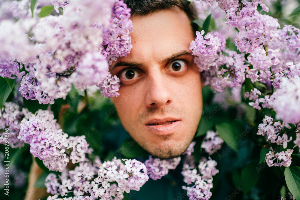 Obraz premium Closeup odd unusual male portrait outdoor at nature in beautiful lilac bushes. Adult funny man face grimace surrounded by blooming flowers. Emotions and facial expxression. Clowning and fooling.