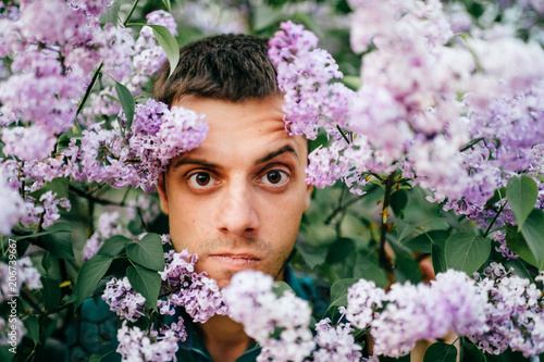 Closeup odd unusual male portrait outdoor at nature in beautiful lilac bushes.  Adult funny man face grimace surrounded by blooming flowers. Emotions and facial expxression. Clowning and fooling.