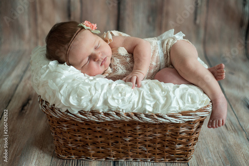 Newborn baby girl in bandage bow sleeping in wicker basket on plaid shawl on beige wooden floor photo