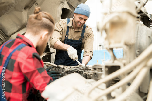 Young professional male mechanic repairing truck in service garage and smiling cheerfully, unrecognizable male colleague helping him