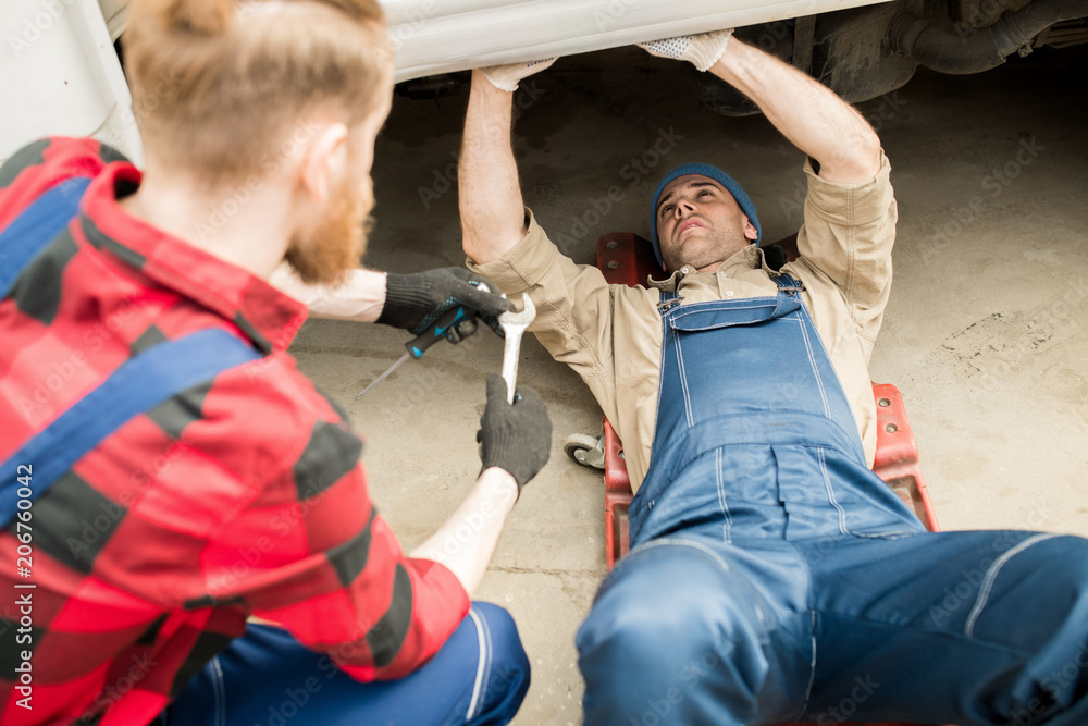 Obraz premium Young professional auto technician lying under truck and fixing breakdown while his colleague consulting him and giving tools
