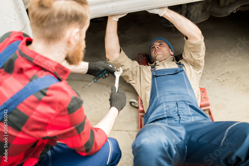 Young professional auto technician lying under truck and fixing breakdown while his colleague consulting him and giving tools