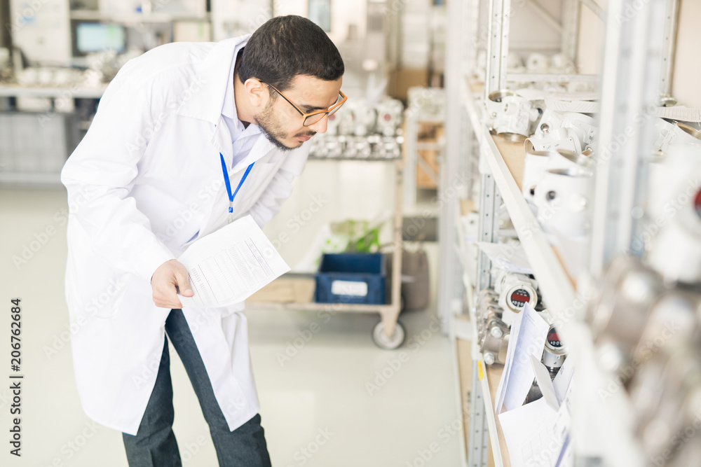 Obraz premium Serious concentrated Arabian engineer in lab coat verifying measuring device list and checking it on shelves in warehouse