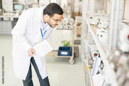 Serious concentrated Arabian engineer in lab coat verifying measuring device list and checking it on shelves in warehouse