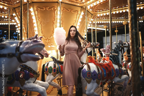 Young beauty model woman posing with old horse carousel in summer park with magic lights