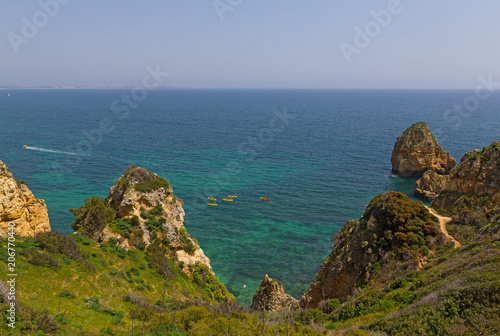 Tourist on kayaks explore grottoes among rocky cliffs along coastline in Algarve, Portugal. Cliffs trail and turquoise sea waters on a warm spring morning.